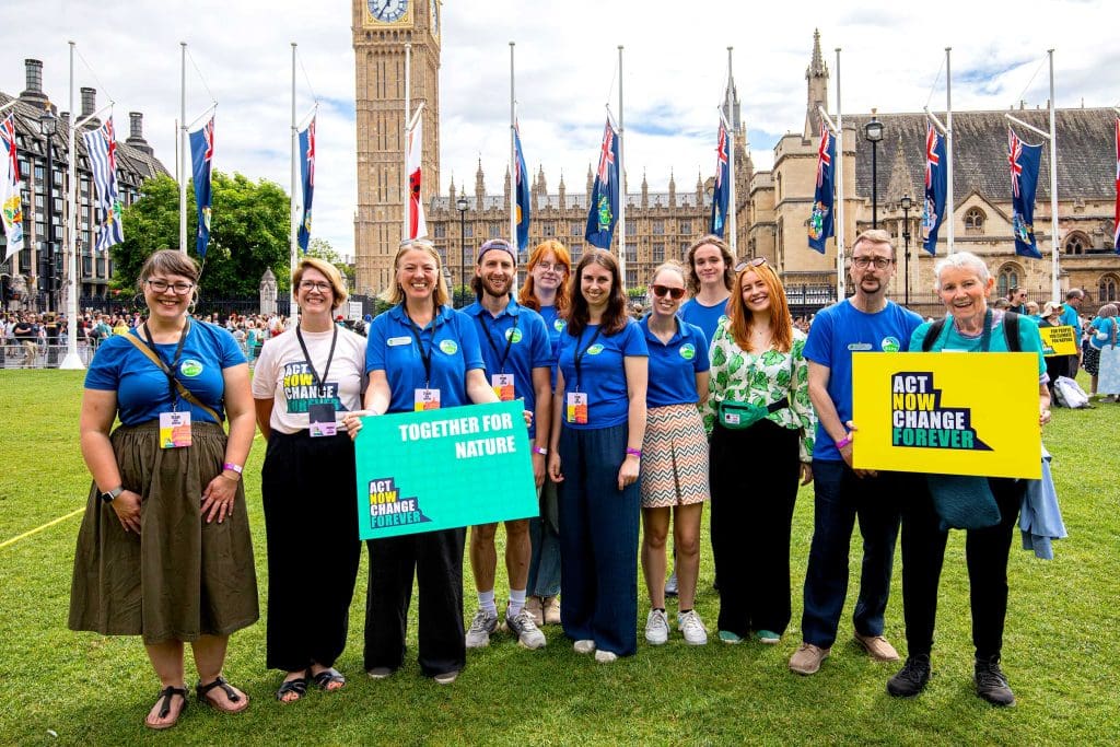 A group of people stand in front of the houses of Parliament holding signs that say 'Act now, change forever' and 'Together for nature'. They are wearing blue RSPB t-shirts