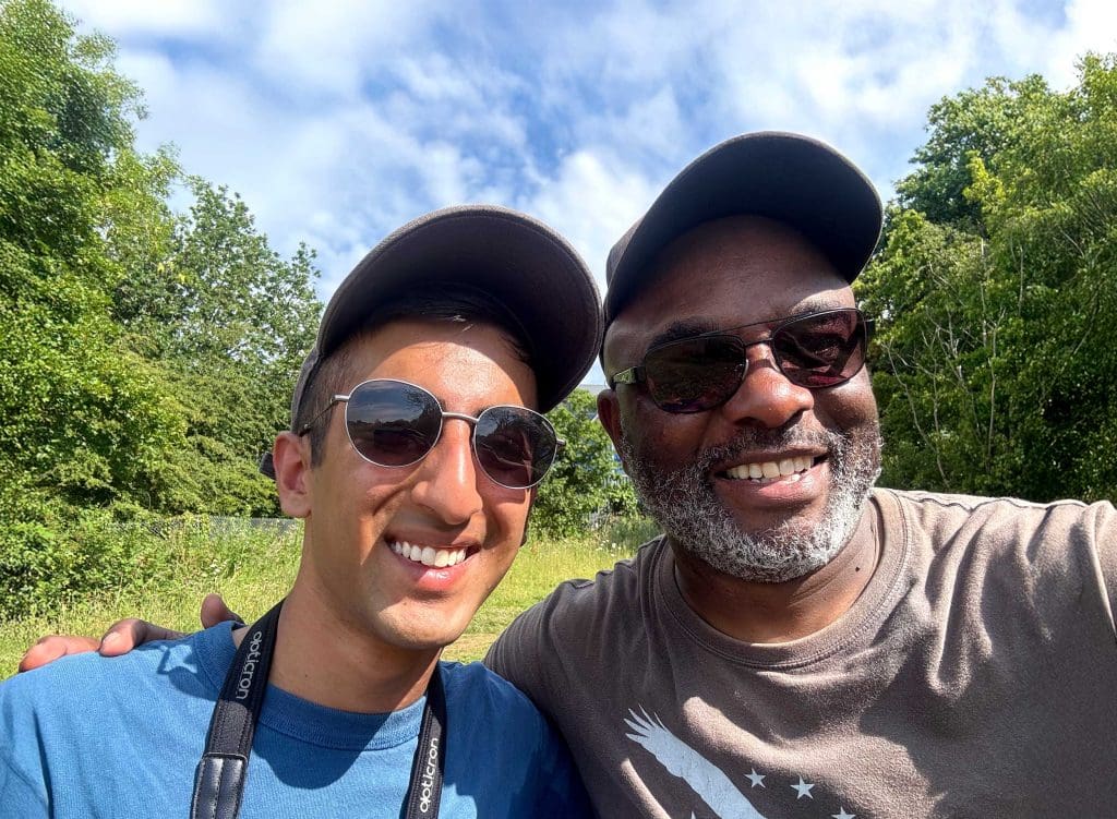 A selfie of Kabir Kaul and David Lindo taken outdoors. Both are wearing sunglasses and caps and smiling at the camera