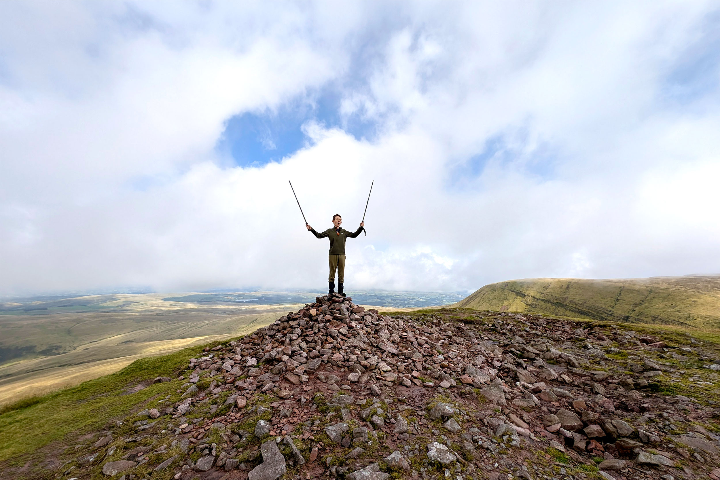 A young boy is stood on a pile of rocks on top of a mountain holding two walking sticks in the air