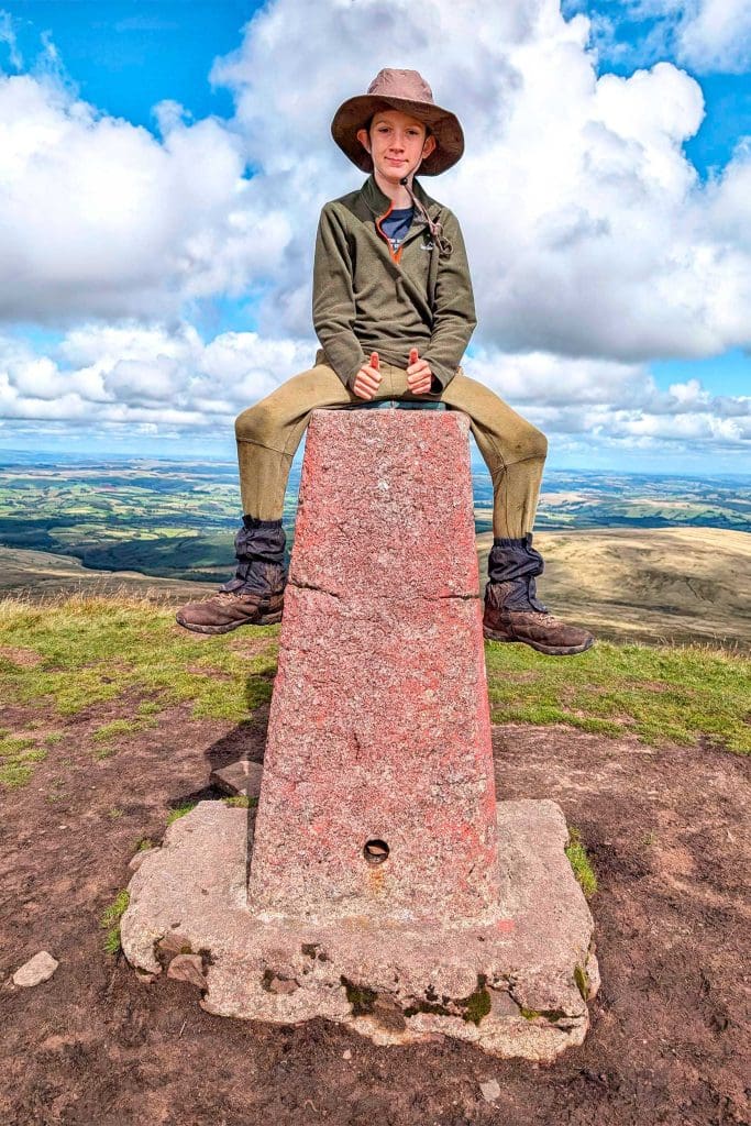 A young boy sits atop a pillar on top of a mountain with his thumbs up