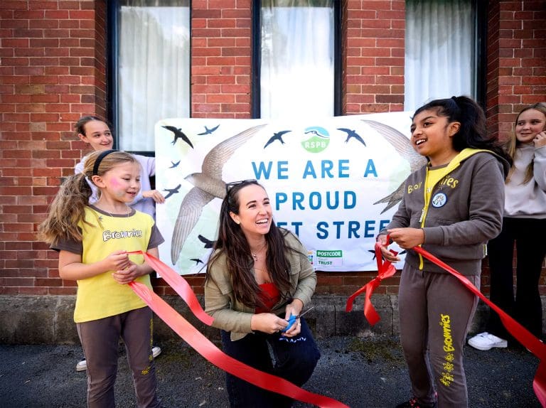 Megan McCubbin with some young Brownies cutting a red ribbon in front of an RSPB sign that reads 'We are a proud Swift Street'