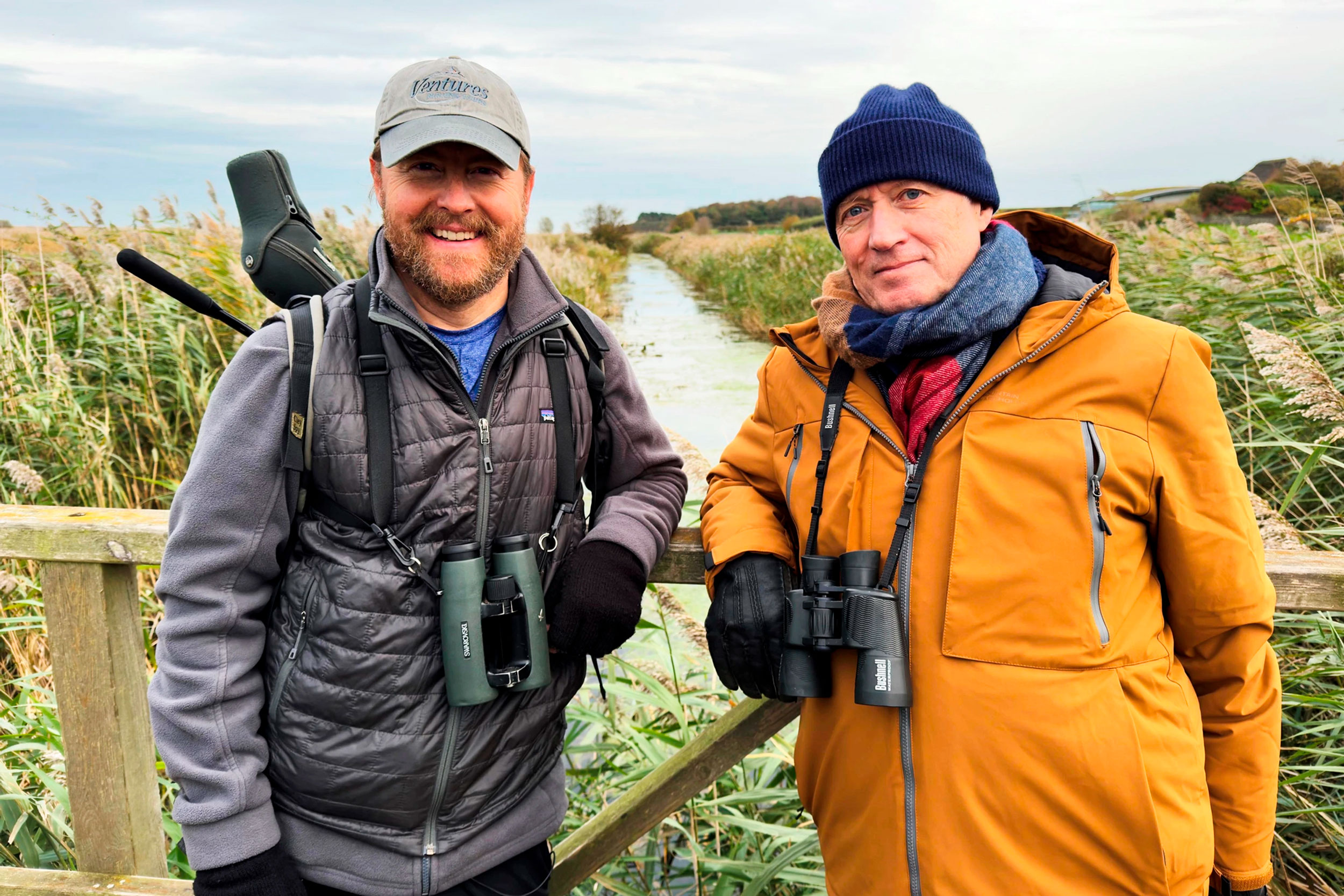 Sam and Ade lean against the side of a small wooden bridge, over a river running through a wetland landscape. They both hold their binoculars