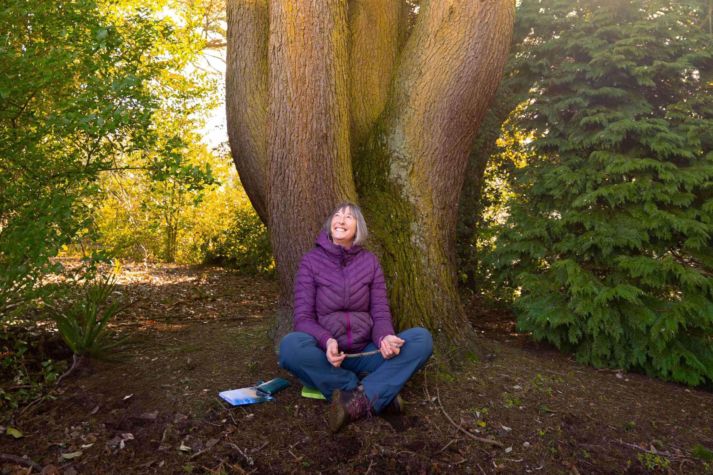 A woman is sitting on the soil at the base of a large tree; we see the trunk only, and surrounding shrubs. She is in the shade, is wearing a coat, and is holding a stick, looking upwards and smiling.