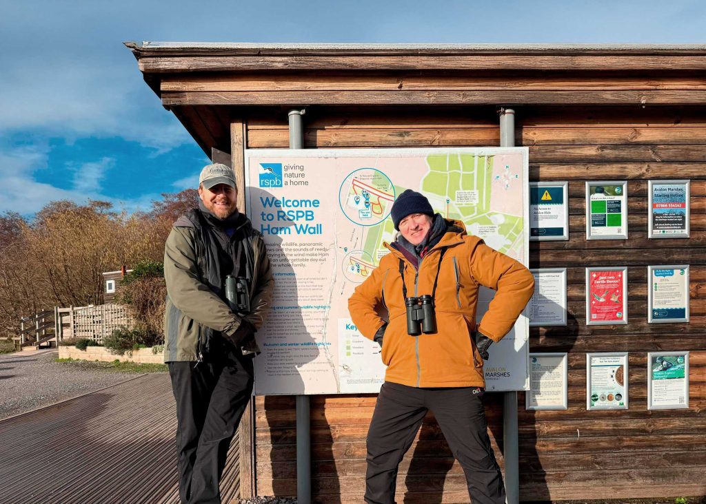 Sam West and Ade Edmondson stand next to an outdoor map of RSPB Ham Wall nature reserve, on a bright, cold day