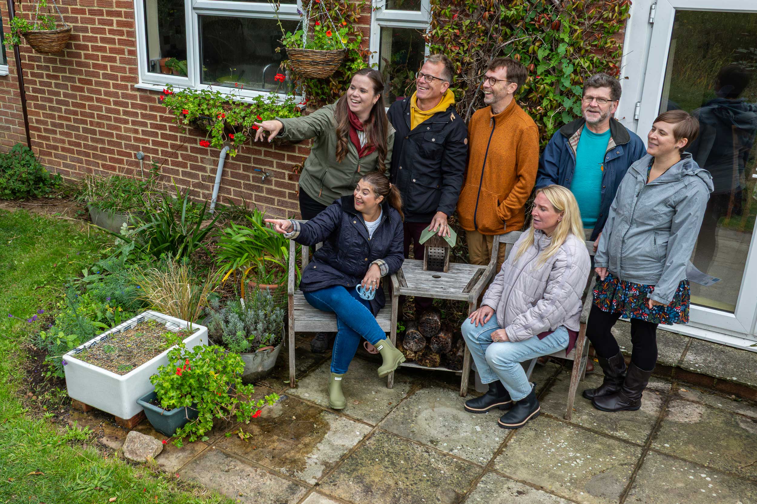 Seven adults, seated and standing, gather in a new build house's back garden to count birds
