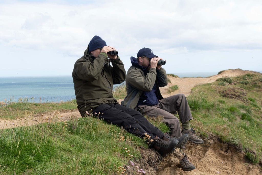 BRDS2-YCST_STILL_1532 Sam and Ade are looking through their binoculars, sitting on the ridge of a grassy trail with the sea in the background