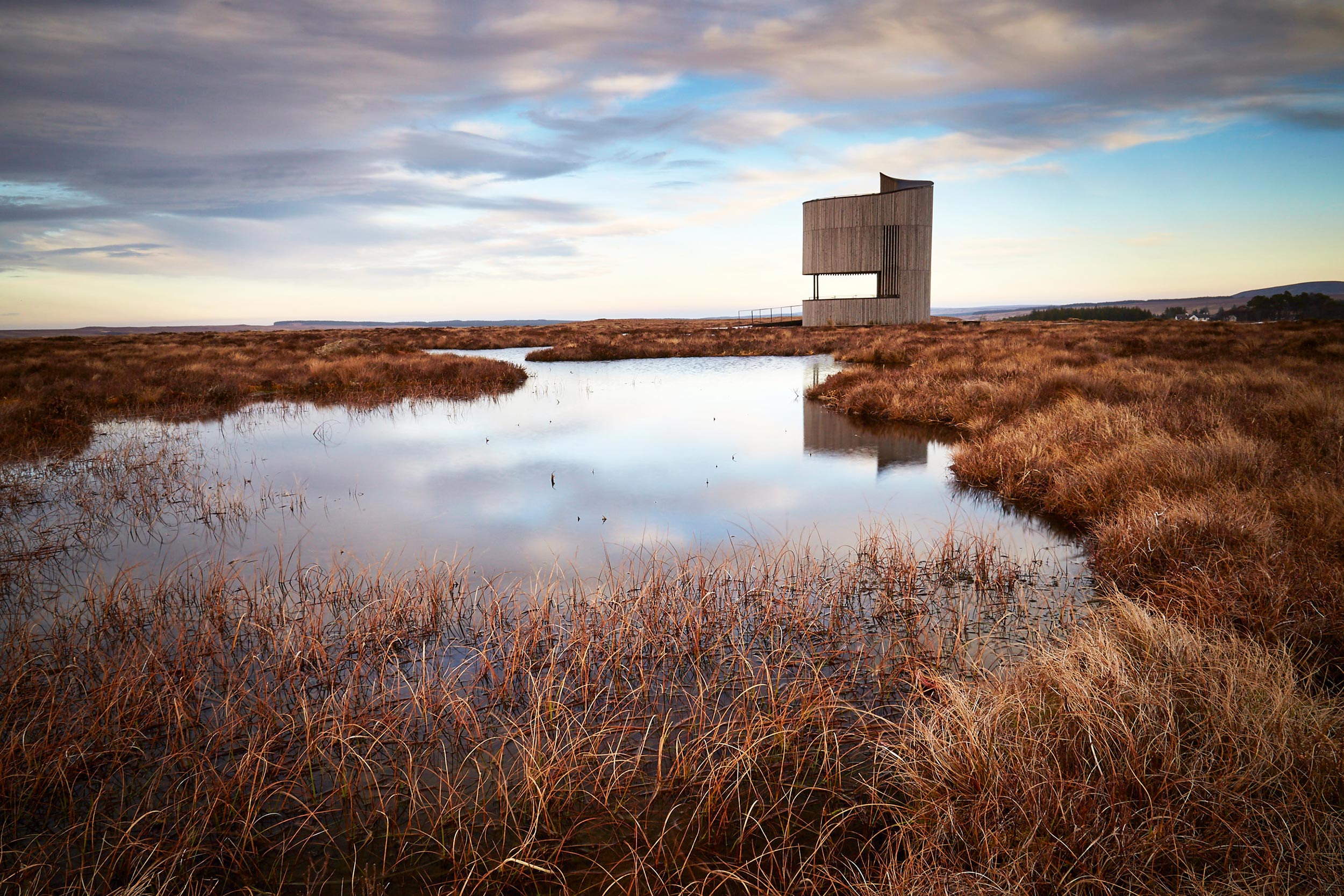 A peatland landscape, with a cloudy sky reflecting in a pool of still water. A tall, birdwatching structure is on the edge of the pool