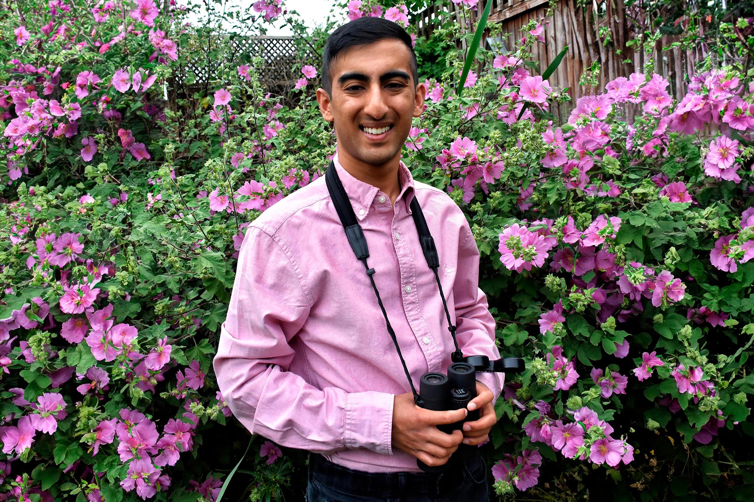 Kabir is standing in front of a pink-flowered bush, wearing a matching pink shirt. he is wearing a pair of binoculars around his neck