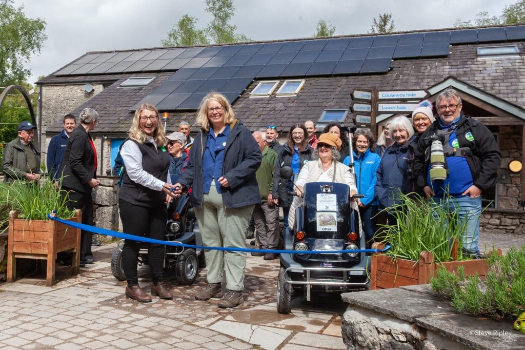 Tramper-Trail-Launch-Event-ribbon1-LM--with-Beccy-Speight-and-Helen-Kirkby---Steve-Ripley A group of people, including two in all-terrain mobility scooters, are gathered behind a blue ribbon which is about to be cut. Behind them is the visitor centre, a low building.